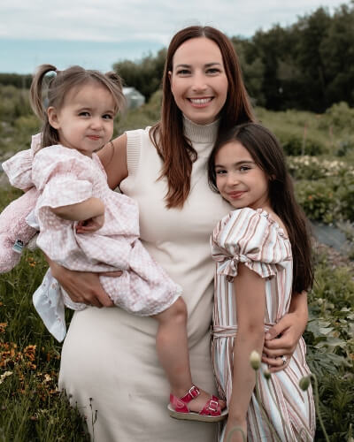 Brittan holding her younger daughter and standing with her arm around her older daughter outside with a forest of evergreen trees in the distance