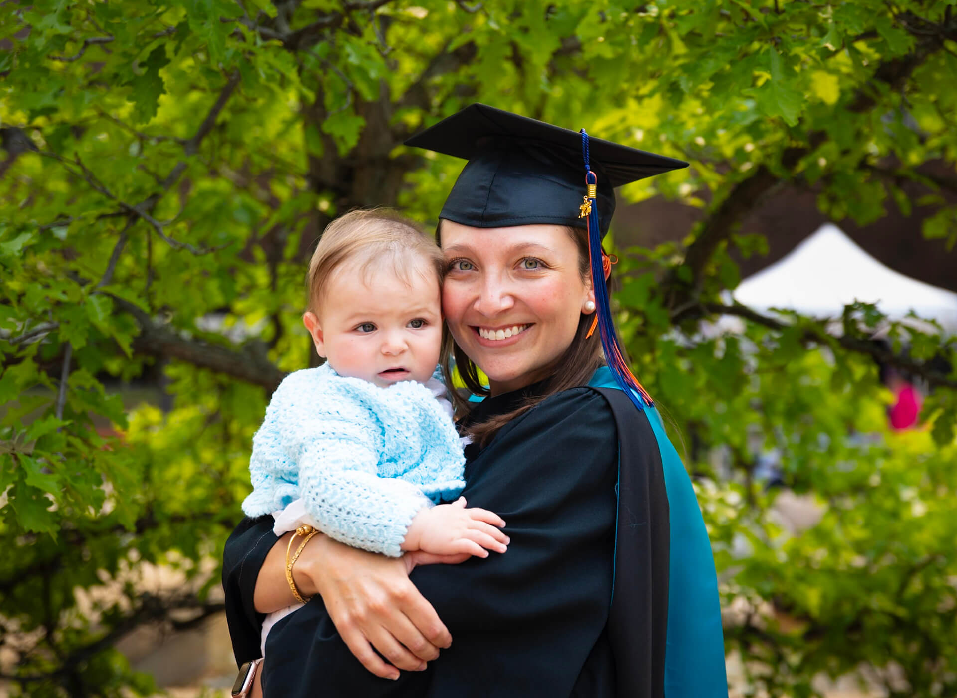 a grad and mom holds her baby with trees in the background