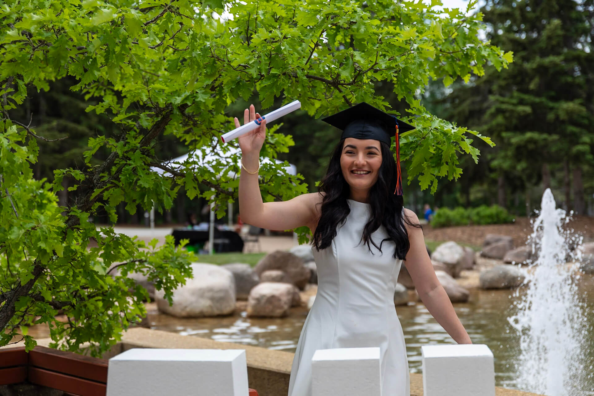 A grad holds their degree in front of a fountain on AU campus