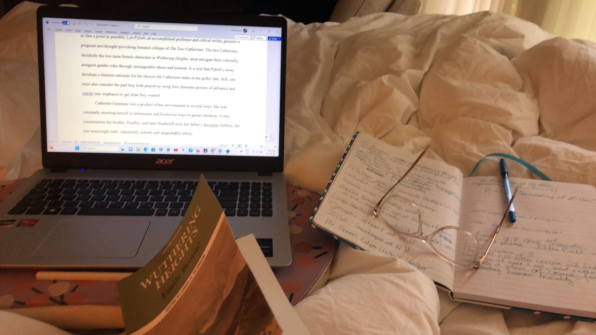 An open laptop along with the book "Wuthering Heights" beside an open notebook with a pen and glasses on a white bedspread