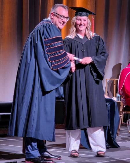 Cori Francis on stage at AU Convocation shaking hands and posing for a photo with President Dr. Alex Clark 