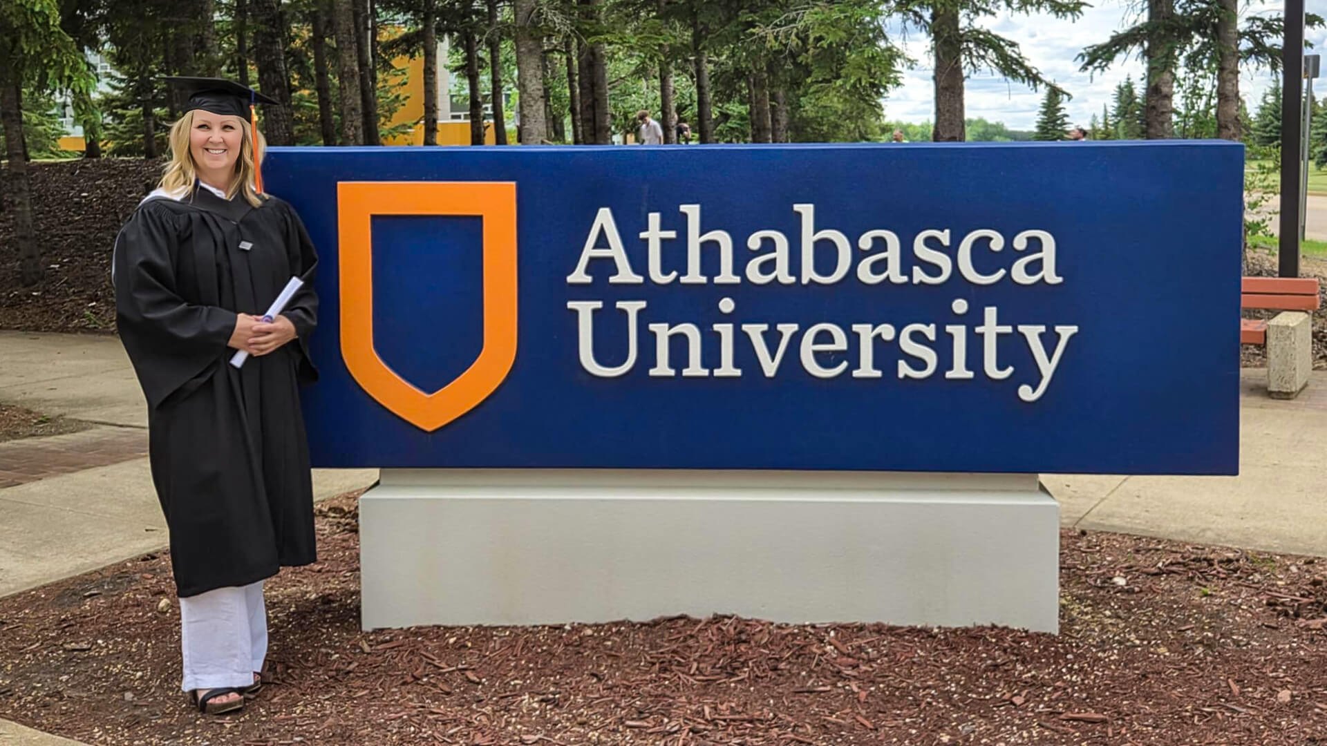 Cori Francis wearing a black cap and gown holding her degree and standing beside the blue Athabasca University sign with an orange sheild.