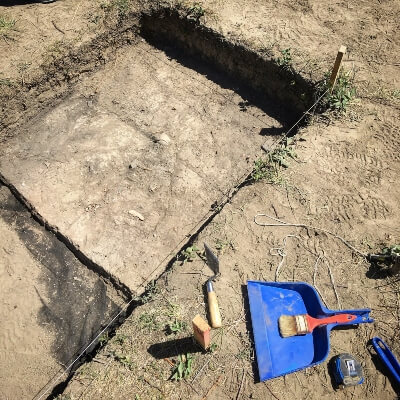 Plot of ground marked off for an archeological dig, with a dustpan, brush and other hand tools on the ground