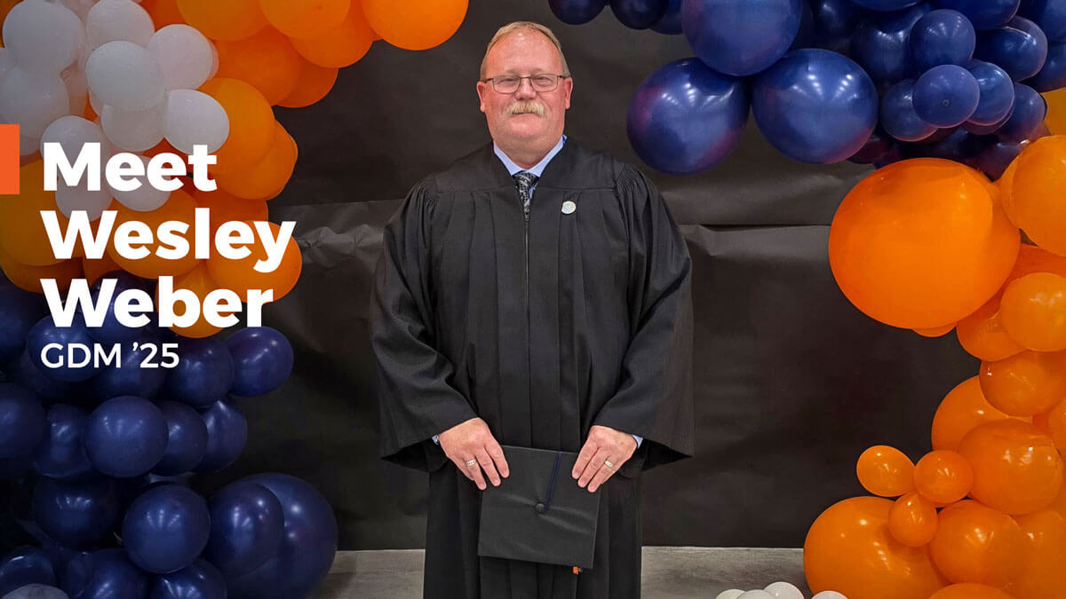 Wesley Weber under an orange and blue balloon arch wearing convocation robe.