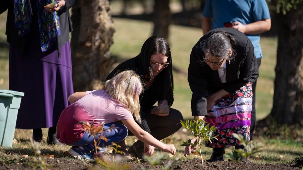 An Elder wearing a colourful ribbob skirt, a woman, and a young girl crouch together in a garden. Two people are behind them, watching as they touch the soil near a plant.