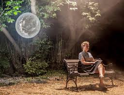 Woman sitting on a bench in a forest setting with a small moon floating behind where she sits