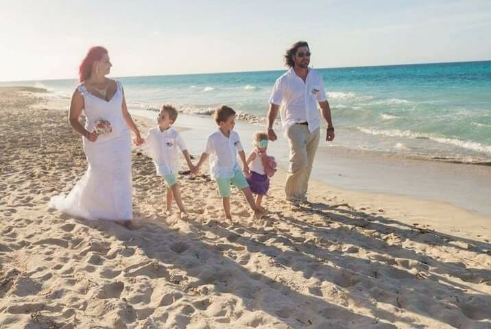 Megan Maksym on the beach wearing a wedding dress holding the hand of one her sons' who's holding hands with his brother, who's holding hands with his sister, who's hands with Megan's husband who is wearing a white colloared t-shirt and khaki pants