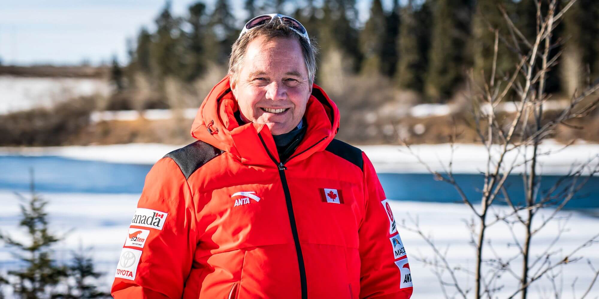 Todd Allison wearing a bright red winter jacket standing infront of a frozen river with pine trees behind it