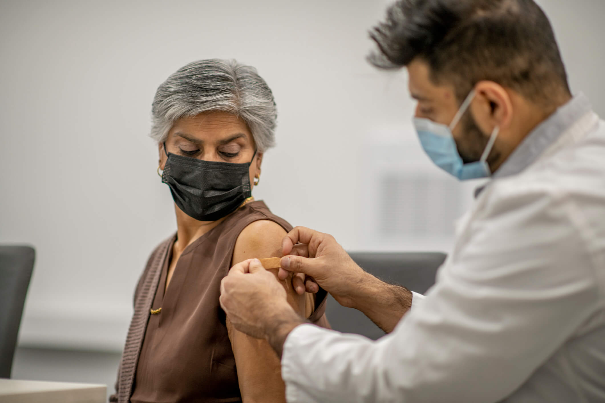 A male doctor puts a band aid on a senior woman's arm after he administered the COVID-19 vaccine injection. They are both wearing a protective face mask to protect themselves from the transfer of germs.