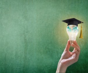 A hand holding up a lightbulb that has a graduation cap on top of it