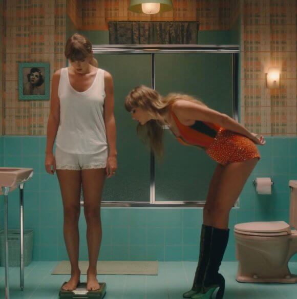 Image of two women (both Taylor Swift) in a bathroom. One of the Taylors is standing on a scale and looking down, the other one is bent over and appears to be peering over at what the scale says
