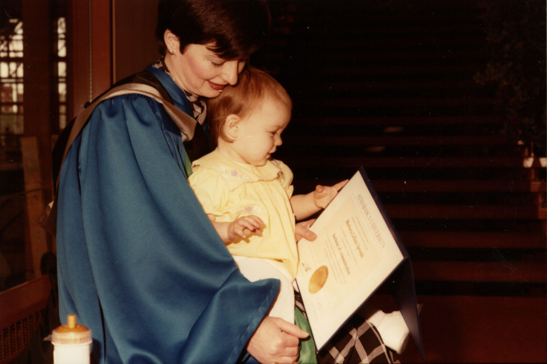 A graduate, presumably a mother, holds a baby and a degree parchment following convocation ceremonies circa 1984
