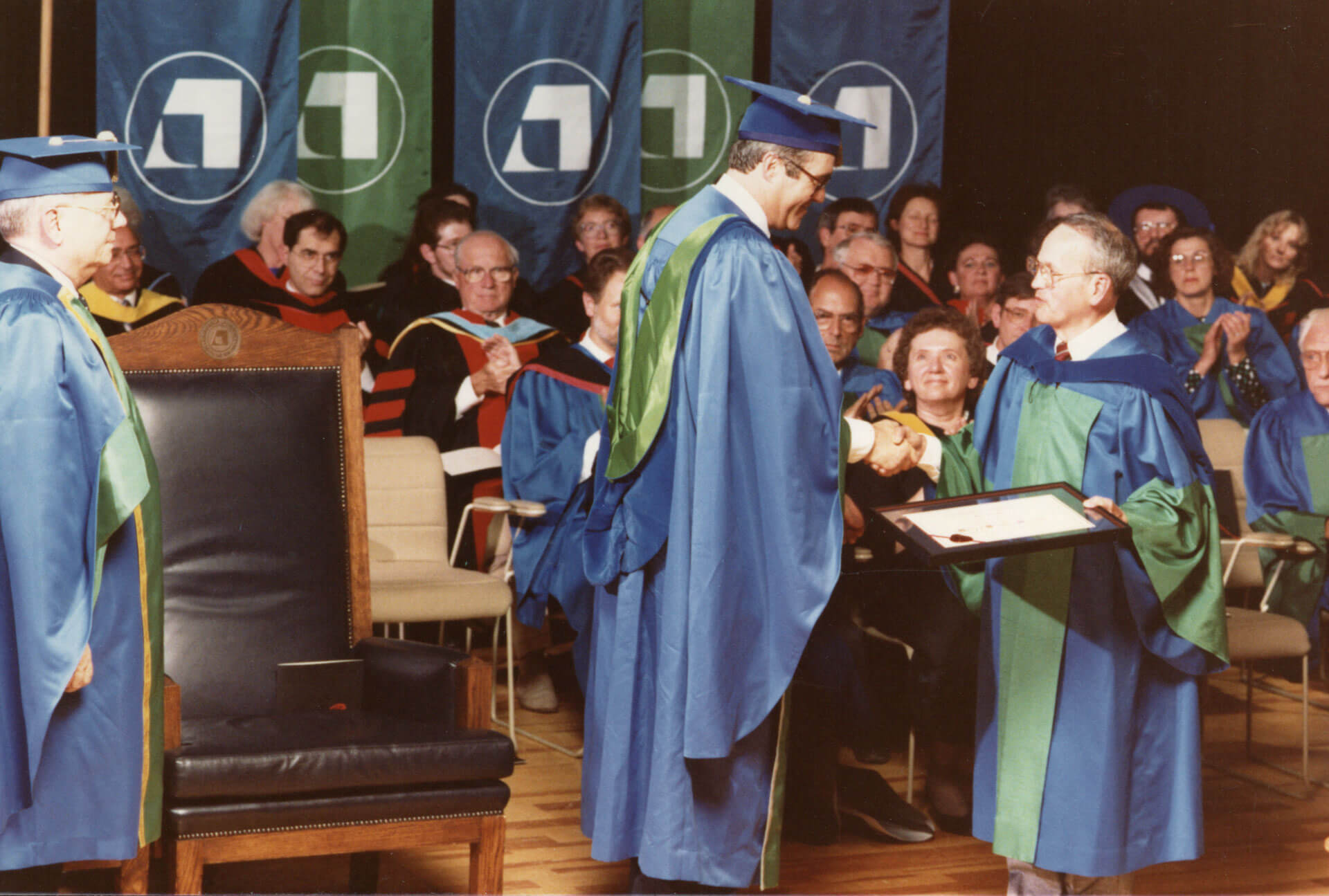 Image of graduate recieiving degree. AU’s founding colours were blue and green, as shown here in this photo circa 1984. Though AU’s brand colours are orange and dark blue currently, the original blue and green can still be seen on AU’s coat of arms. The colours reference AU’s connection to central and northern Alberta.