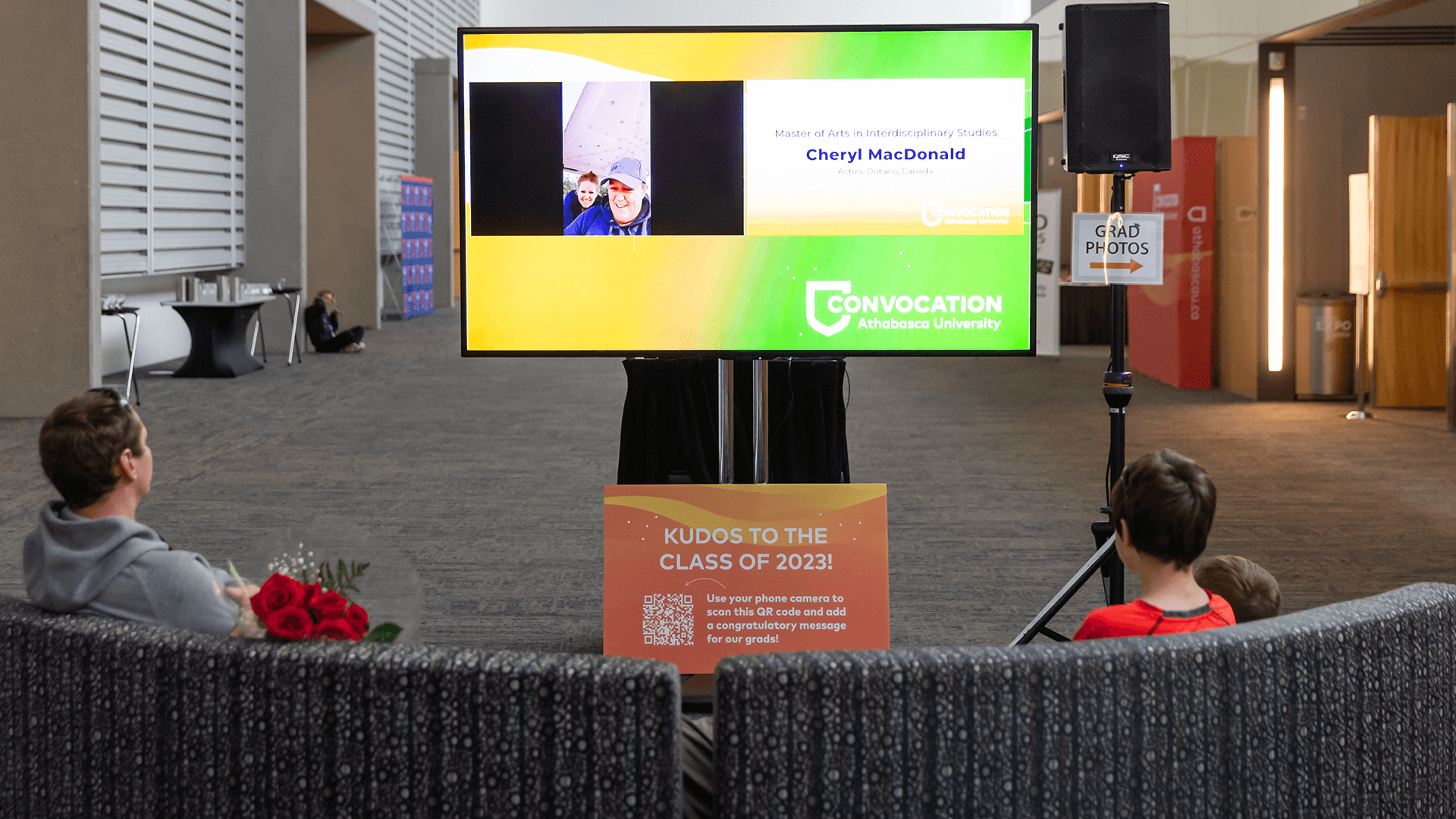 image of two people sitting on a couch outside the ceremony space looking at a stream on a big television screen of the convocation ceremony