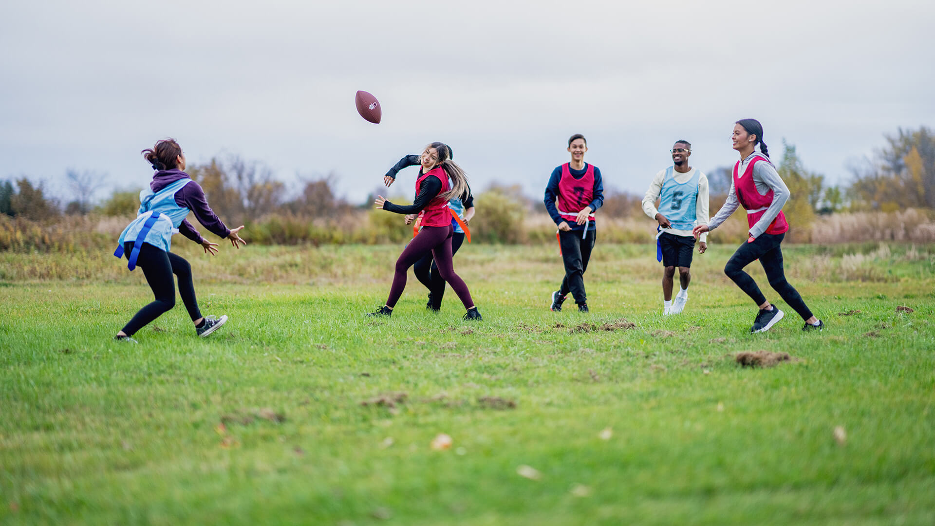 A group of University students run around on a field as they play a game of flag football together. They are each dressed comfortably and have numbered team pinnies on.