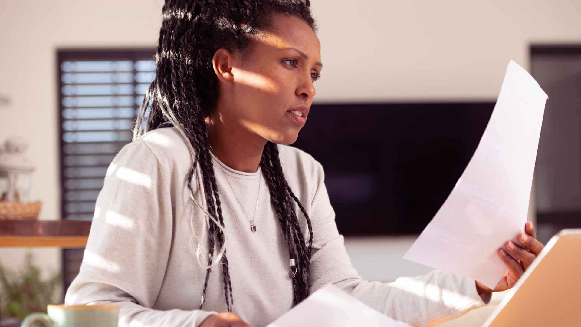 woman-reading-documents-home-office-research-study-routinestrategy Person reading a document while working on a laptop in a well-lit office space.